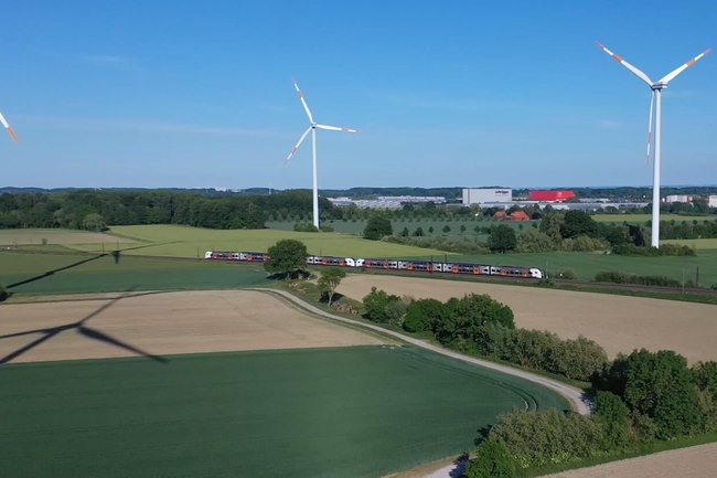 Regionalzug fährt durch eine unbebaute Landschaft, im Hintergrund Windräder