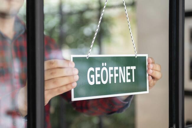 Ein Mann öffnet sein Geschäft. Auf einem grünen Schild steht "Geöffnet"