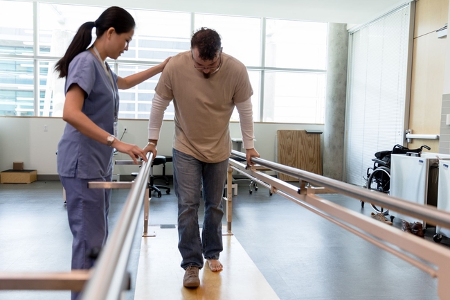 Patient in der Rehabilitation übt zu gehen und wird dabei von einer Pflegefachfrau betreut