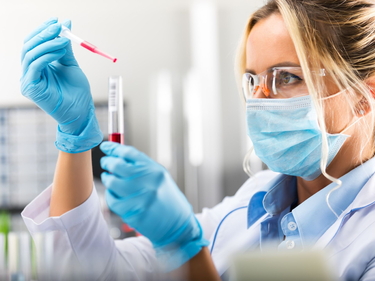 Laborantin mit Maske und Schutzbrille hält Reagenzglas mit Arzneimittel in der Hand